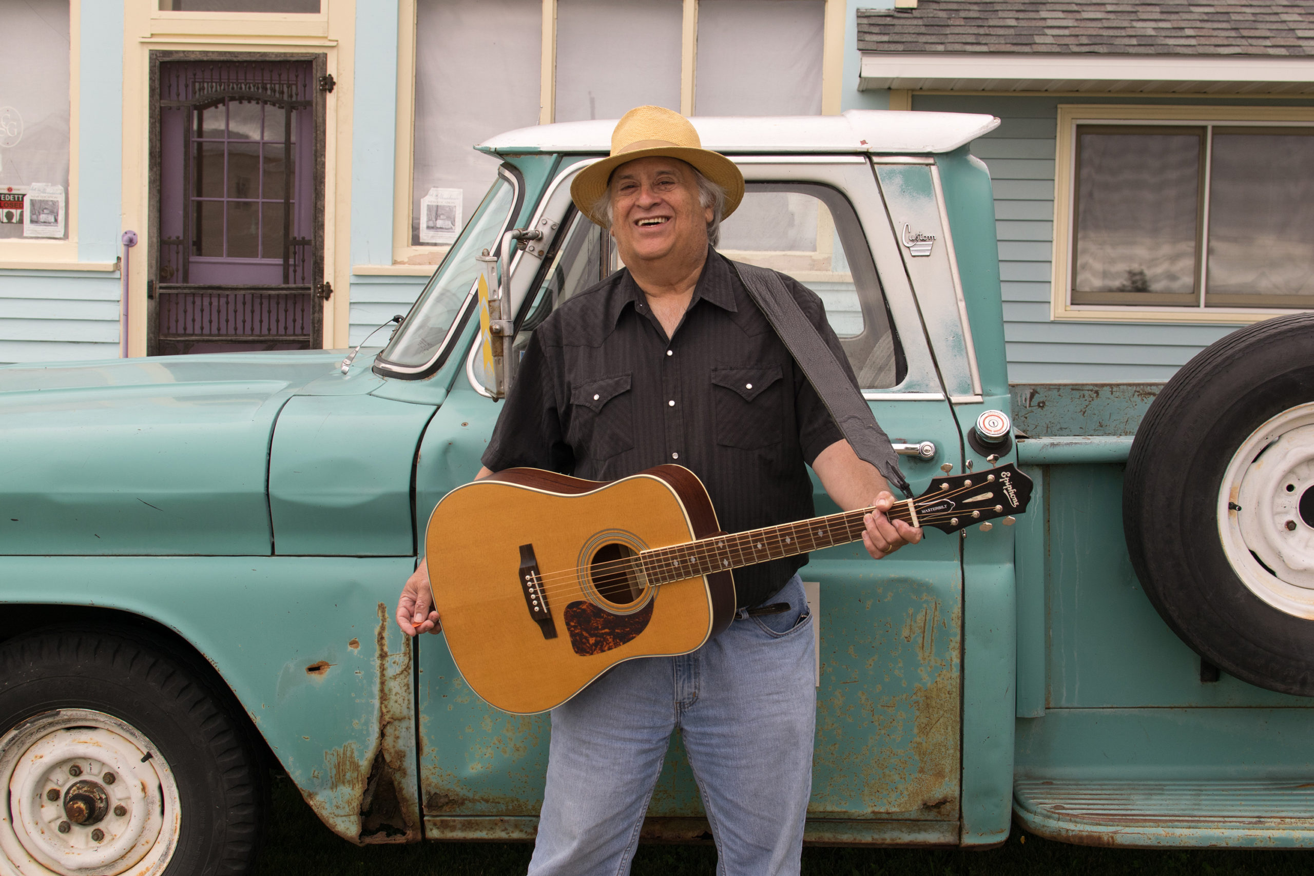 Man with acoustic guitar leaning on a baby blue pick up truck.