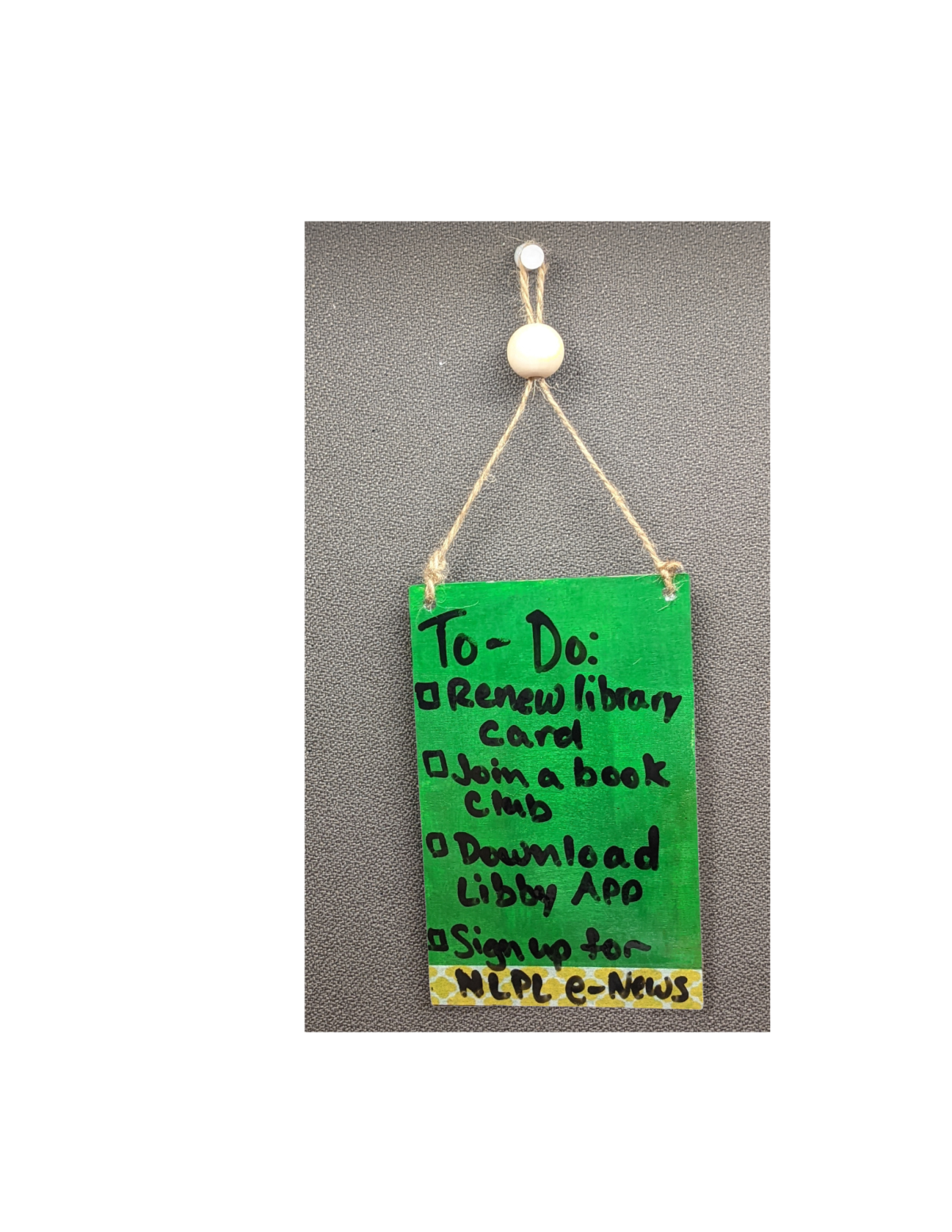 Green doorhanger with a string hanging from a push pin. Text reads " To-Do: Renew Library Card, Join a Book Club, Download Libby App, Sign up for NLPL e-News"